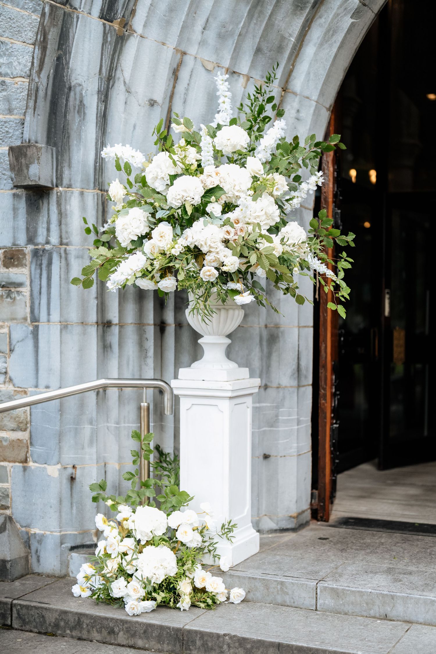 Elegant white floral arrangements in urns decorate a stone church entrance with arched doorways.