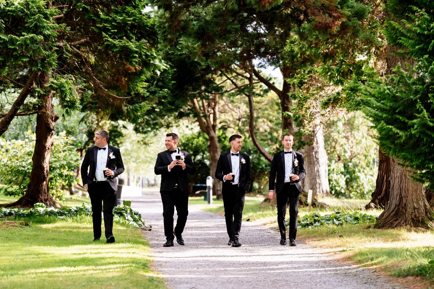 Wedding party wearing formal black attire walks along tree-lined path in lush garden setting during outdoor celebration.