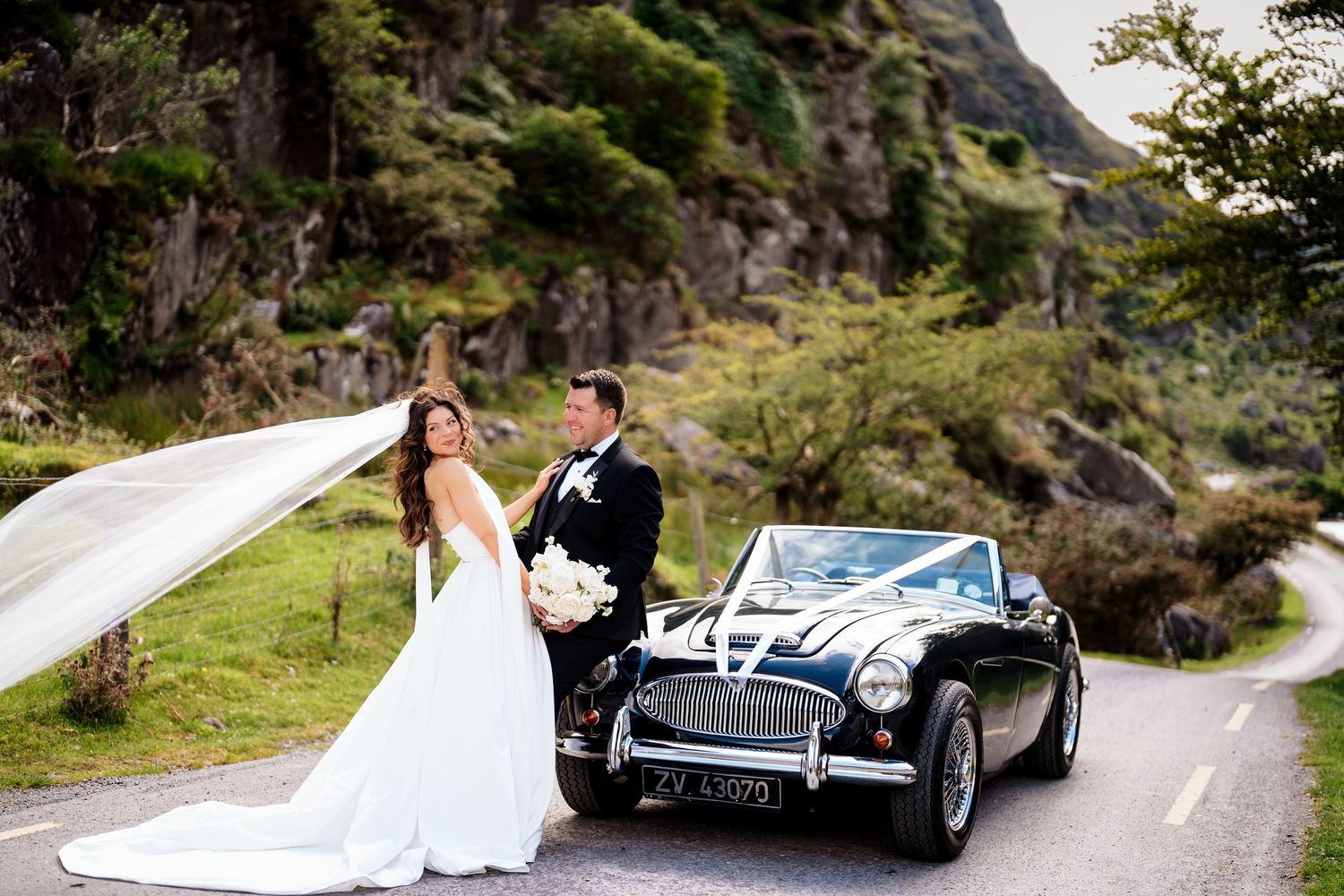 Newlywed couple poses with classic black vintage car on a winding mountain road with flowing white veil.