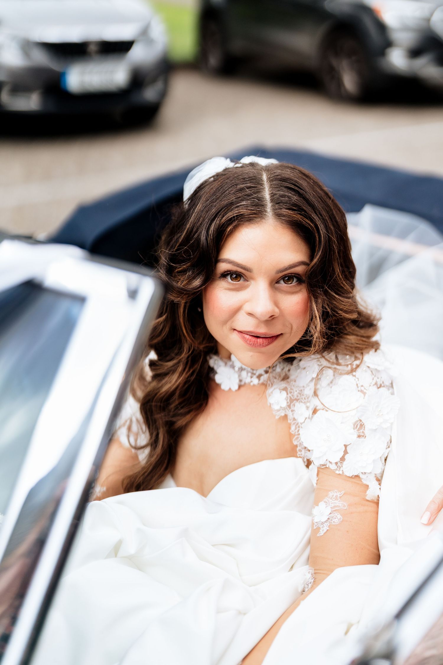 Woman in white lace wedding dress poses elegantly near parked cars outdoors.