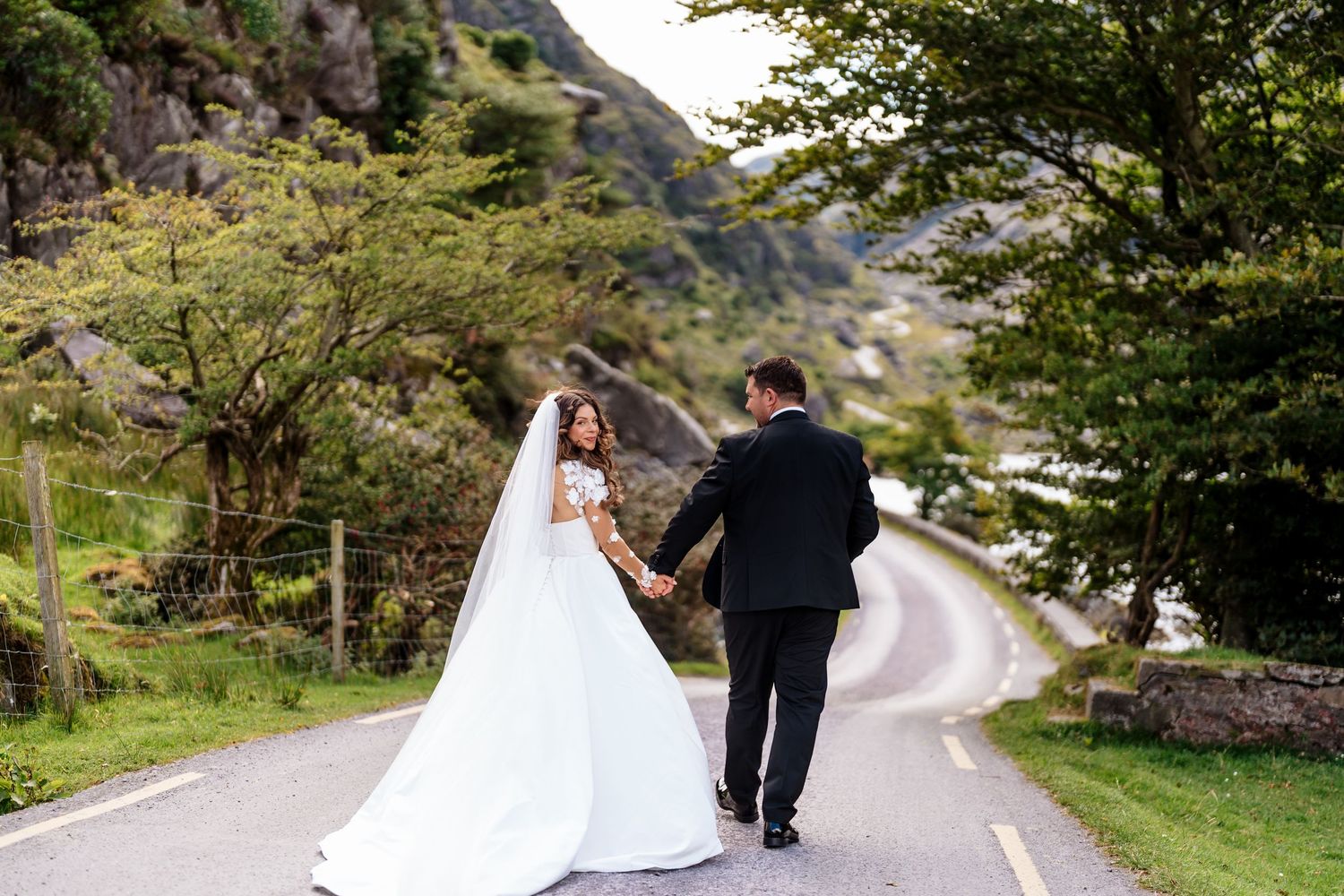 A wedding couple walks along a winding mountain road surrounded by lush green scenery.