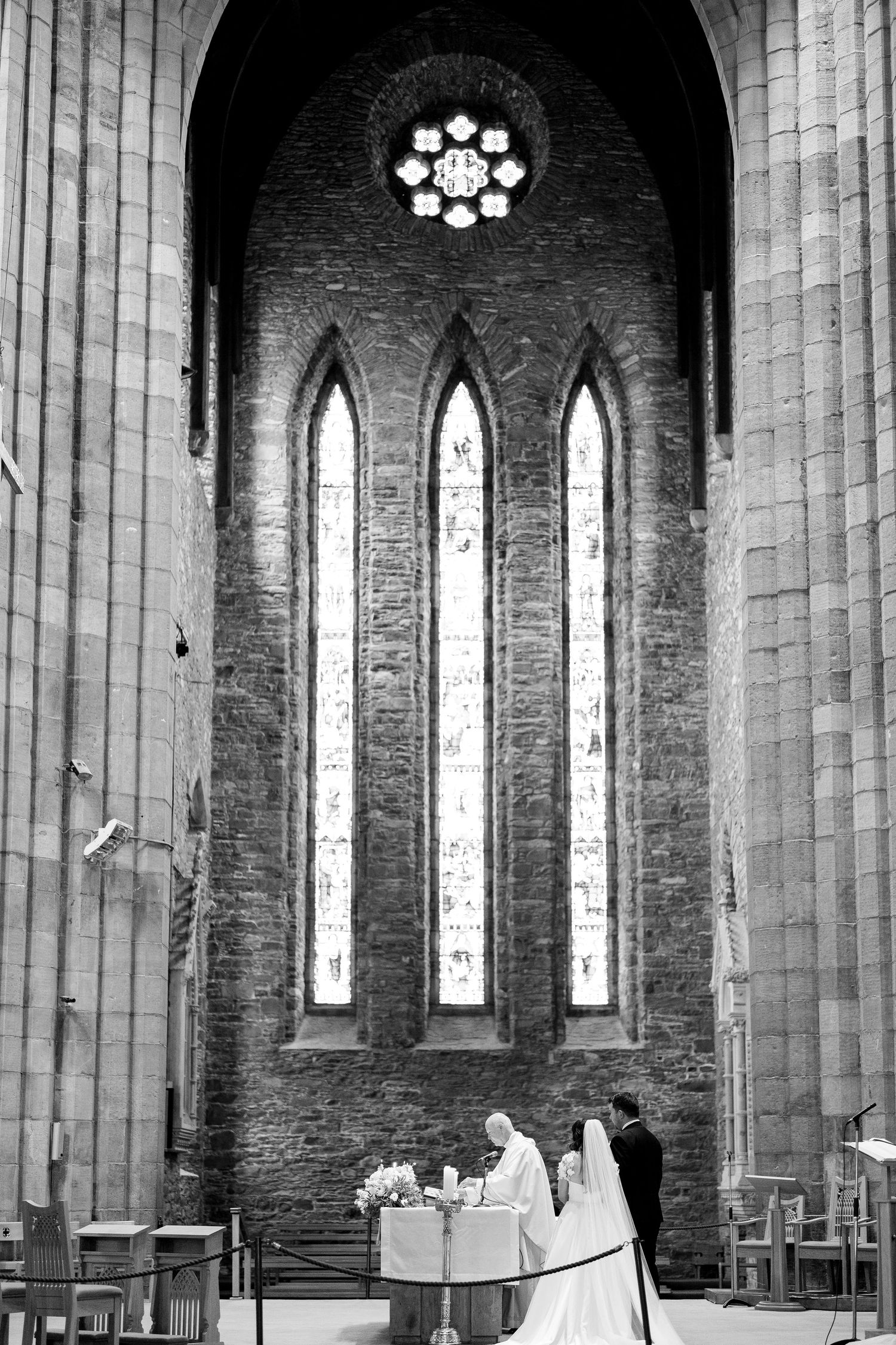 Inside an old stone church with tall arched windows and columns, a wedding ceremony takes place in the grand space.