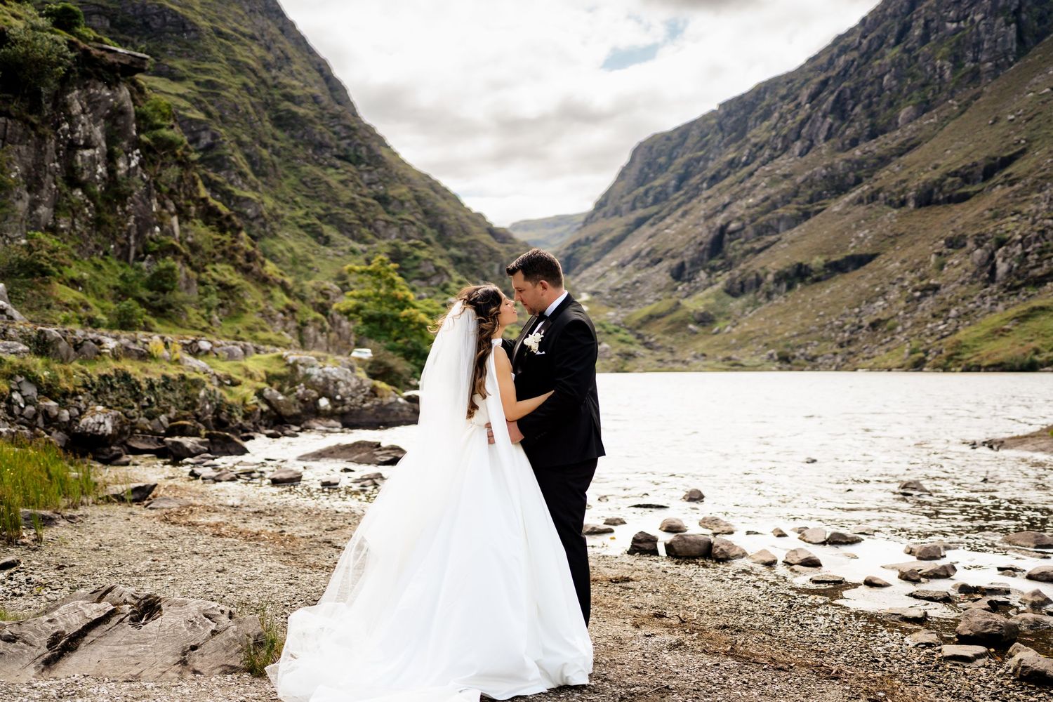 A couple in wedding attire poses in the dramatic Scottish Highlands landscape with mountains and lakes in the background.
