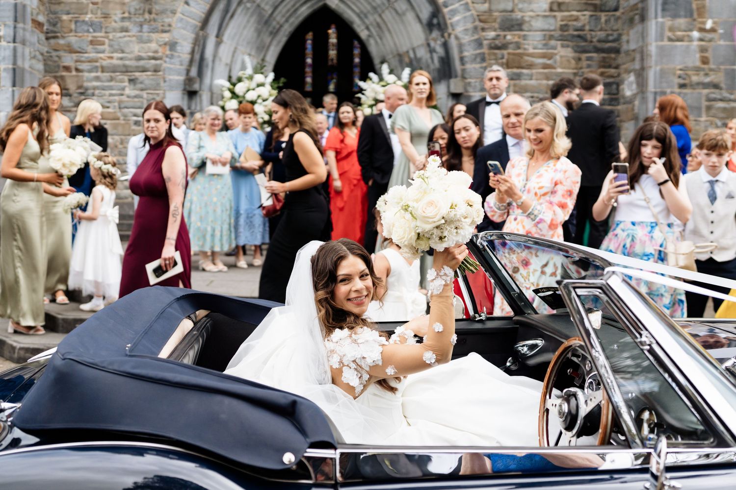 A convertible car drives past a crowd of wedding guests throwing confetti and celebrating outside a stone church.