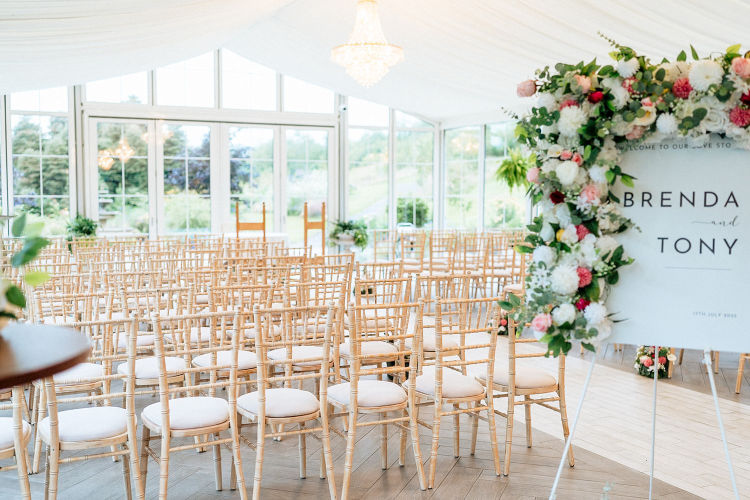 Elegant indoor wedding ceremony setup with wooden chairs and floral arrangements in a bright conservatory space.