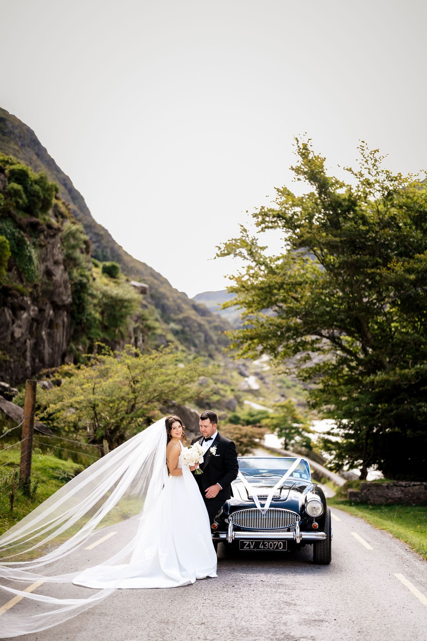 Elegant wedding couple poses with classic vintage car on scenic mountain road surrounded by lush greenery.
