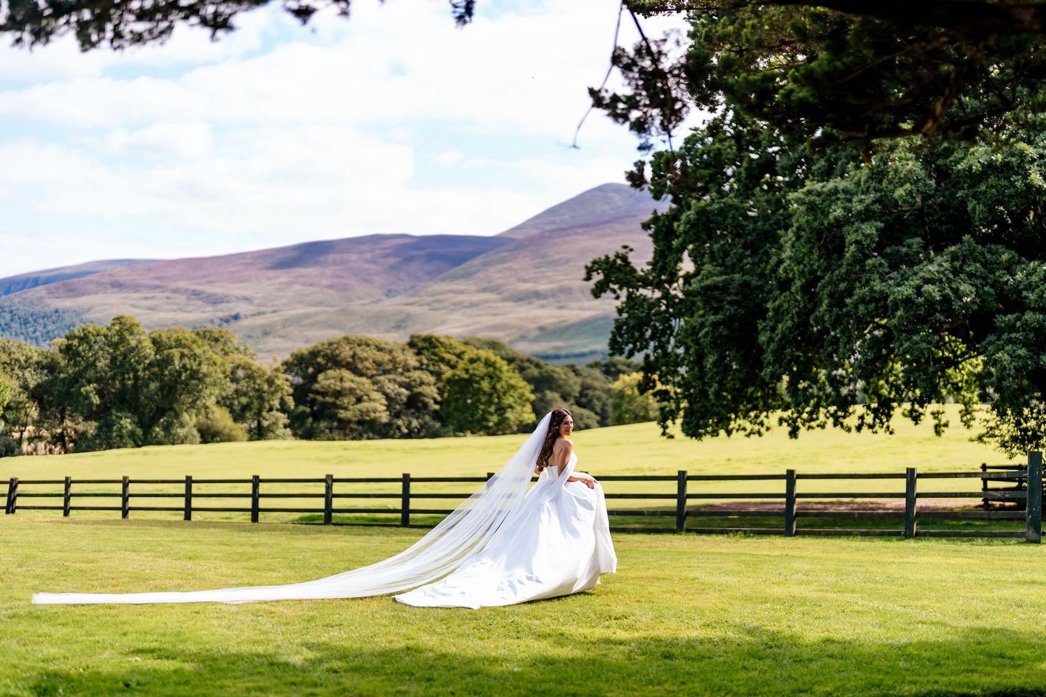 Mountain wedding ceremony at a scenic ranch venue with green fields and wood fencing under a beautiful blue sky.