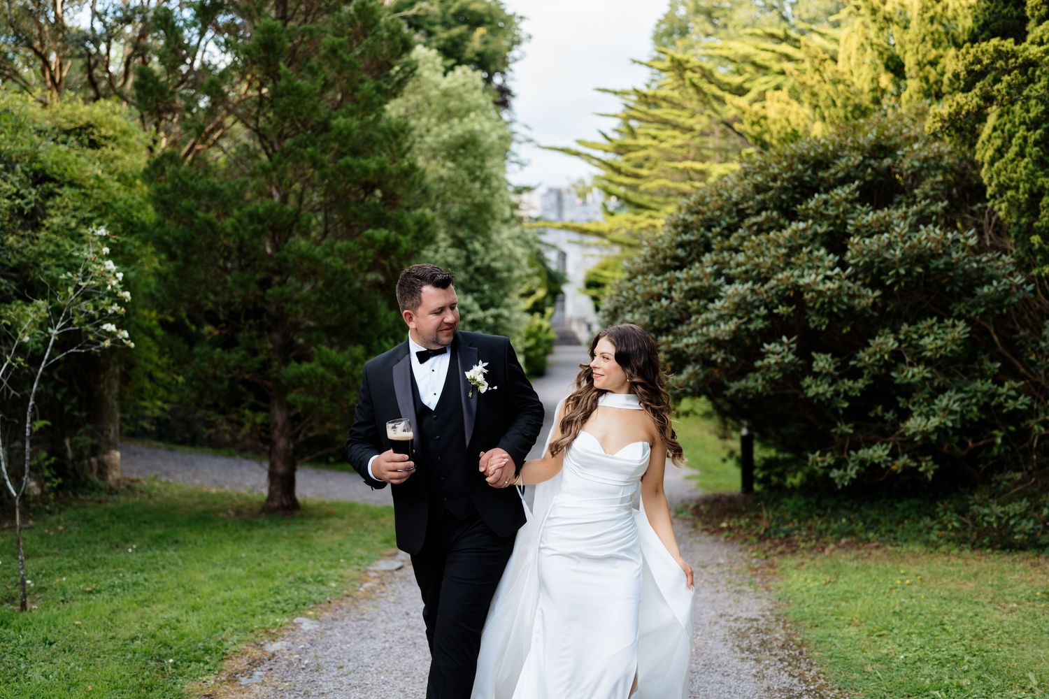 Two people in formal attire walking through a lush garden pathway together on a sunny afternoon.