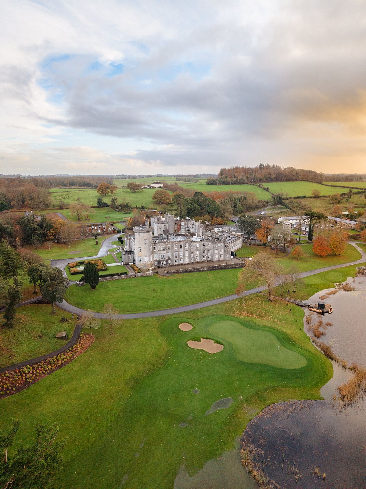 Dromoland Castle Aerial view of a grand castle hotel with golf course and lake surrounded by rolling countryside at sunset.