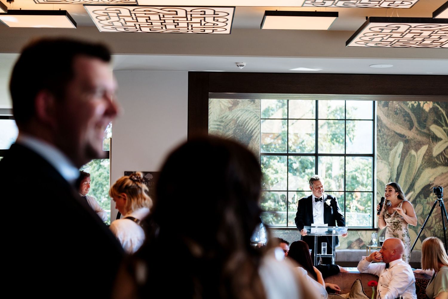 Guests watch and listen attentively during a wedding reception speech in an elegant venue with large windows.