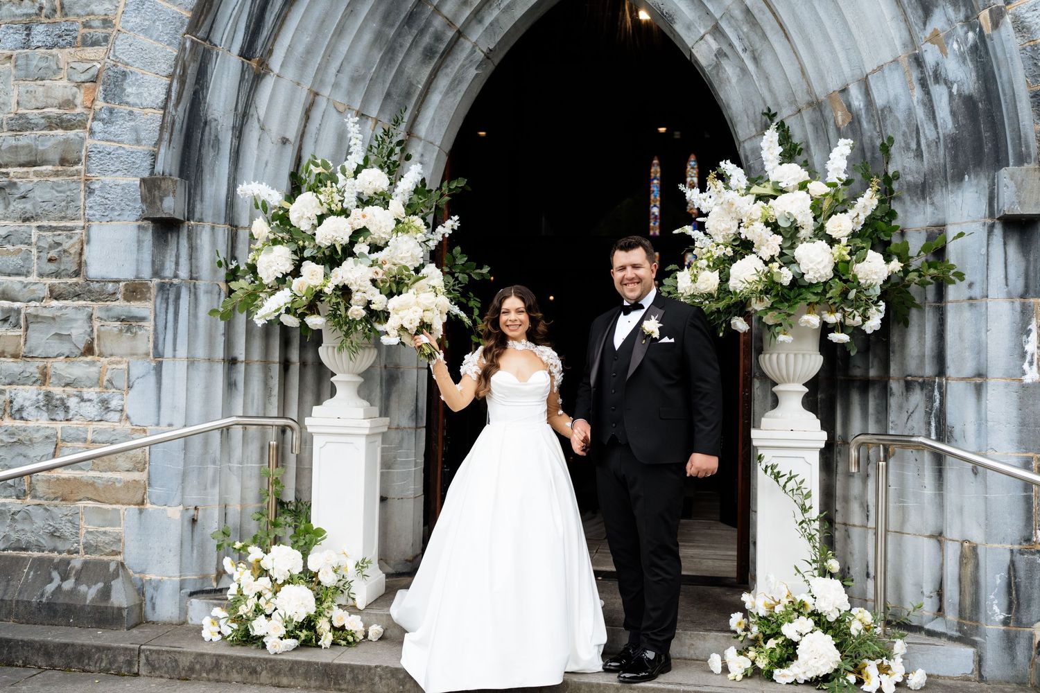 A couple exits a stone church archway decorated with white floral arrangements during their wedding ceremony.