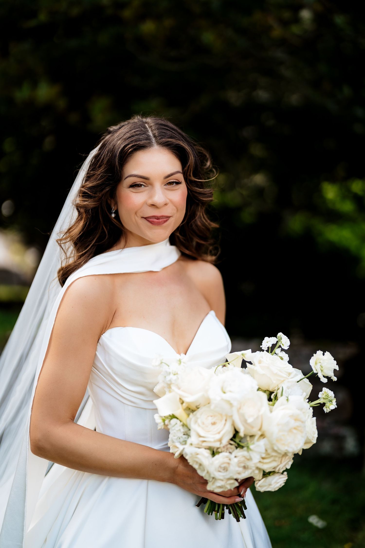 A bride in a white wedding dress holds an elegant bouquet of fresh white roses in an outdoor garden setting.