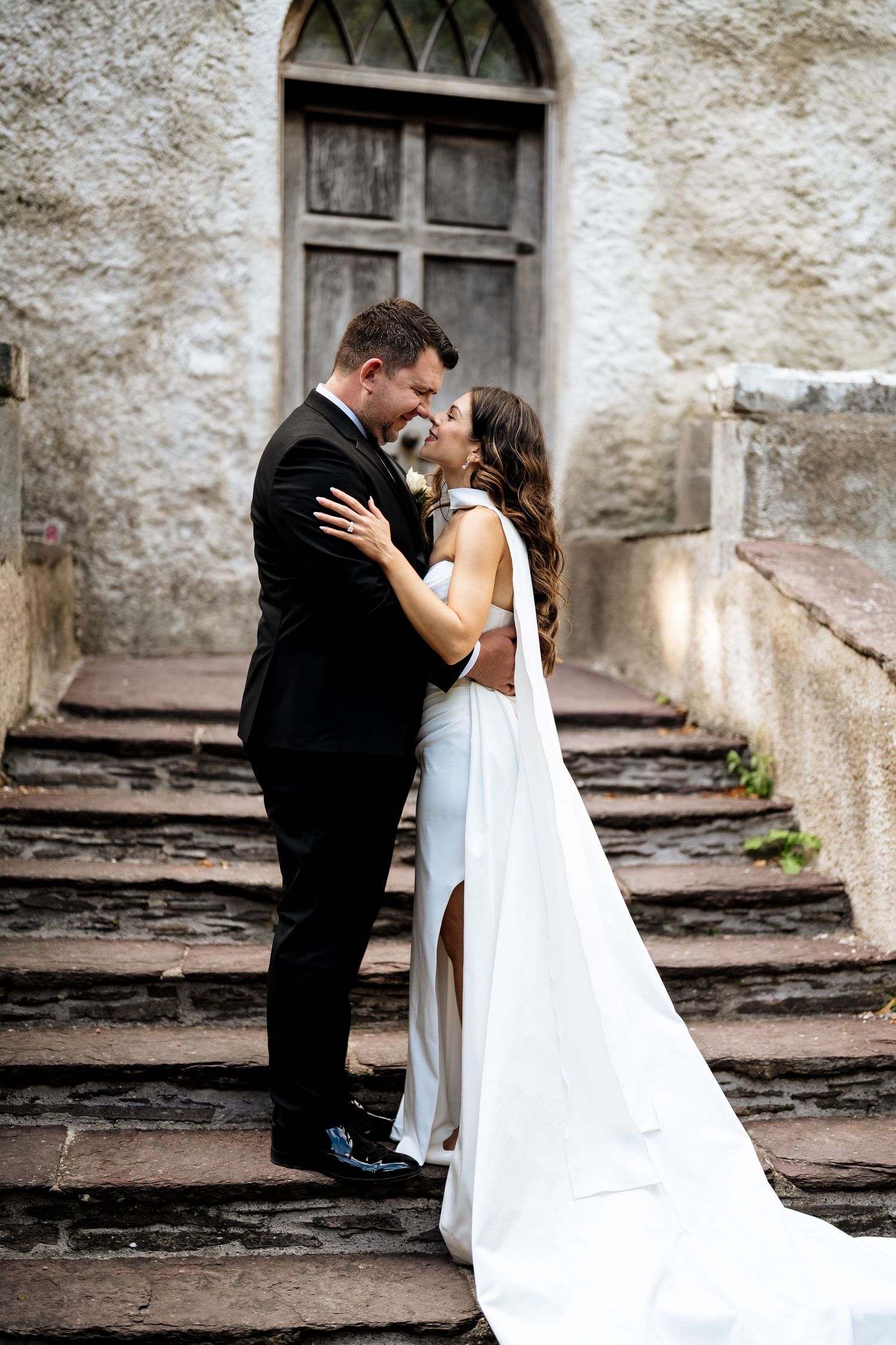 Romantic couple shares intimate moment while seated on historic stone steps in front of weathered building facade.
