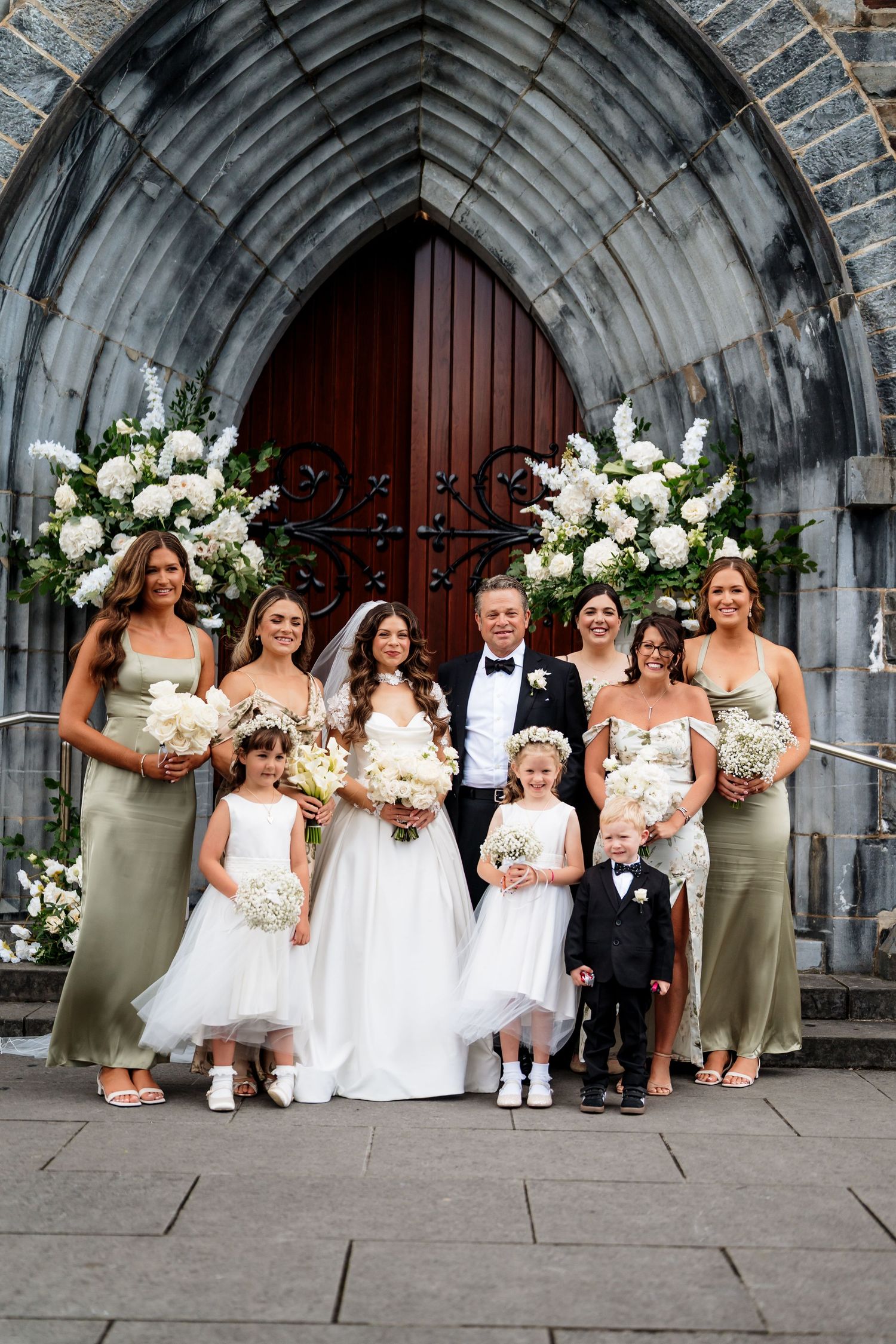 Wedding party poses together under stone archway decorated with white floral arrangements for formal portraits.