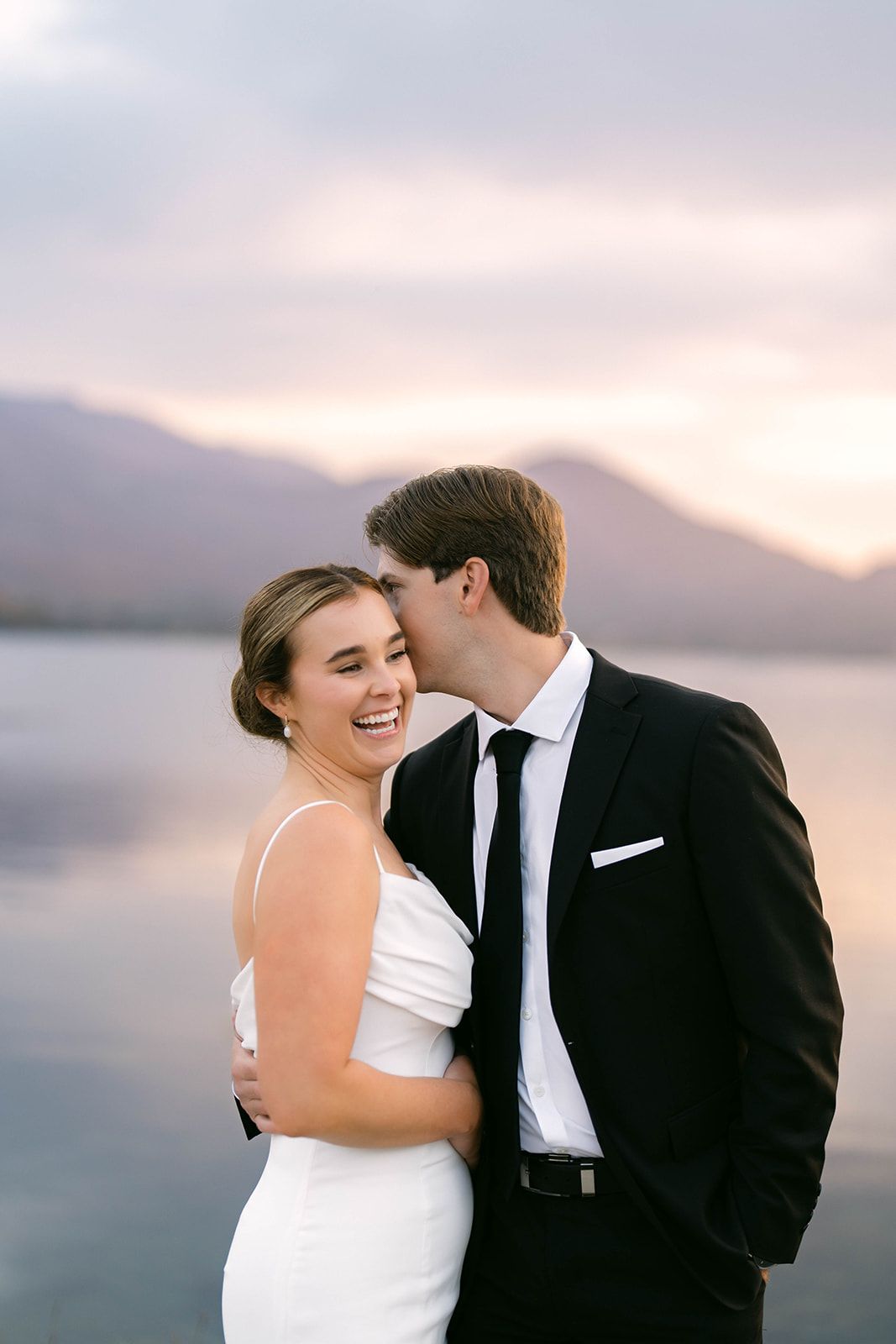 A romantic wedding couple shares an intimate moment at sunset by a serene lake with mountains in the background. Sheen Falls wedding photos