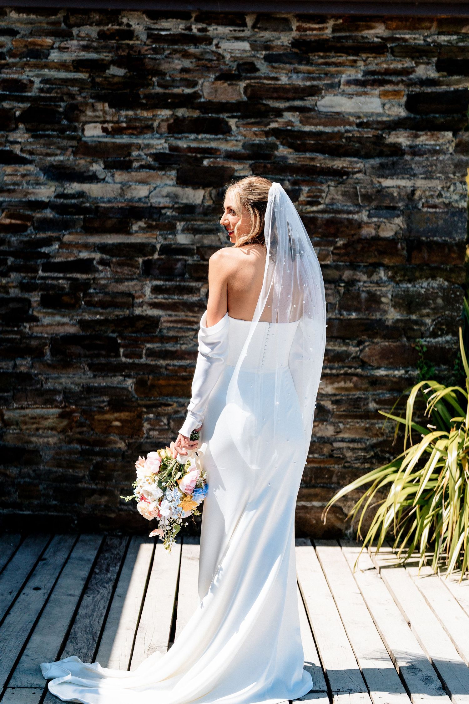 A bride in an elegant white backless dress and long veil holds a pastel bouquet while standing against a stone wall.