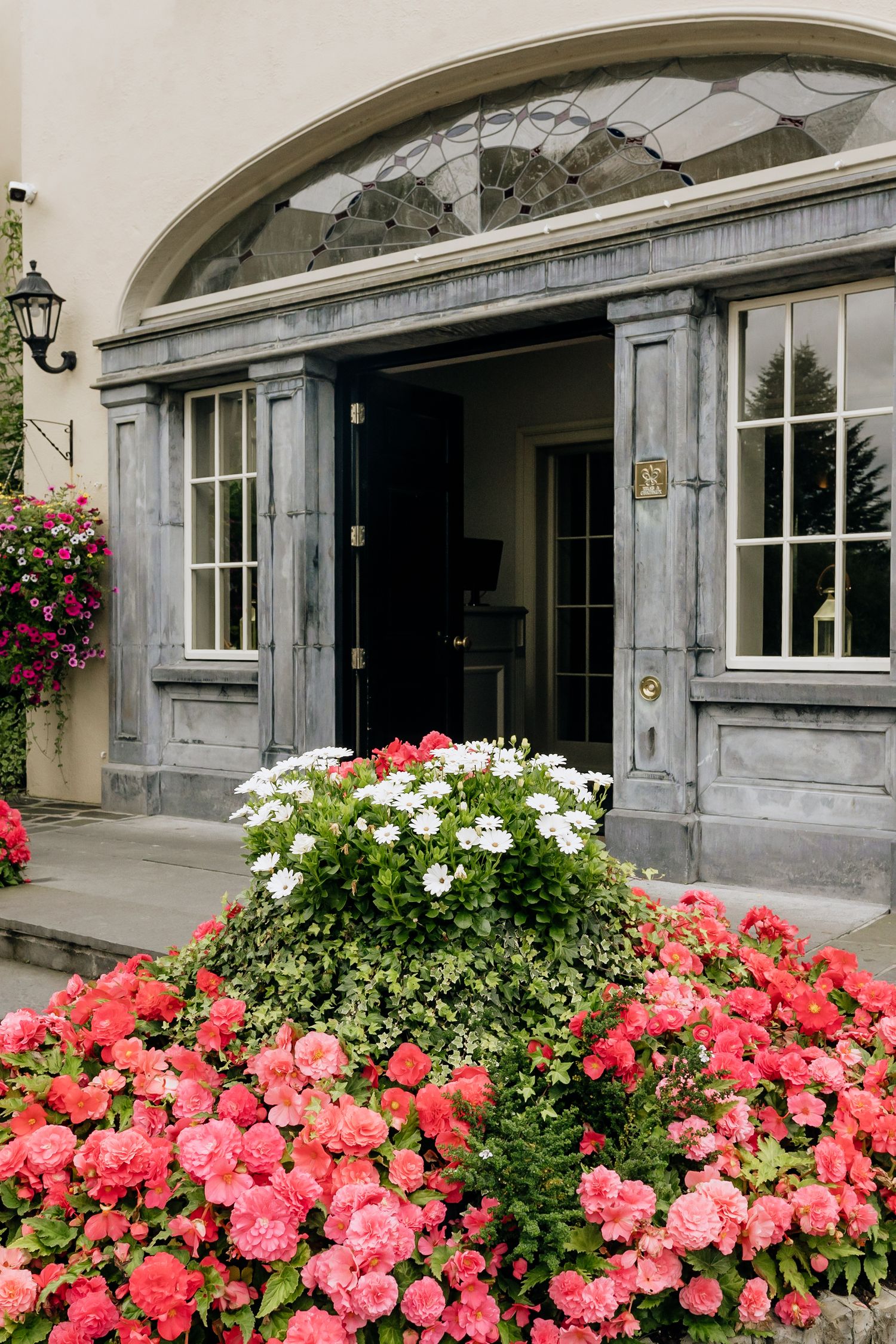 Elegant entrance archway adorned with vibrant pink and white flowers in hanging baskets and garden beds.