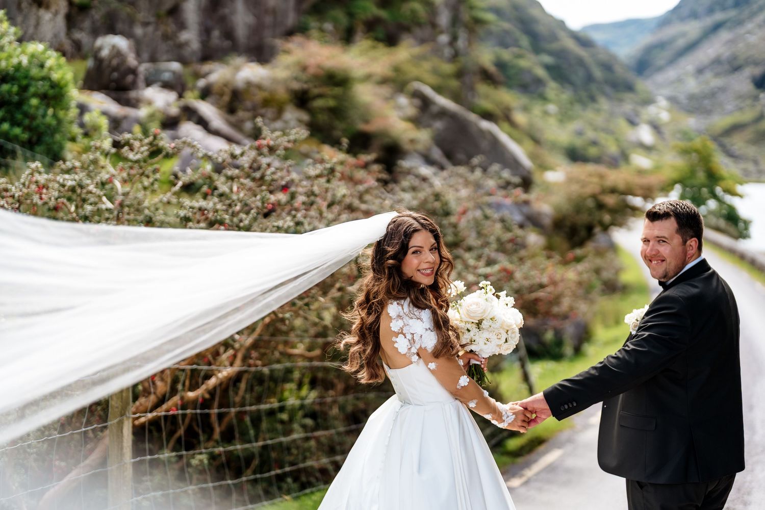 A couple celebrates their wedding day against a scenic mountain backdrop with a flowing white veil catching the wind.