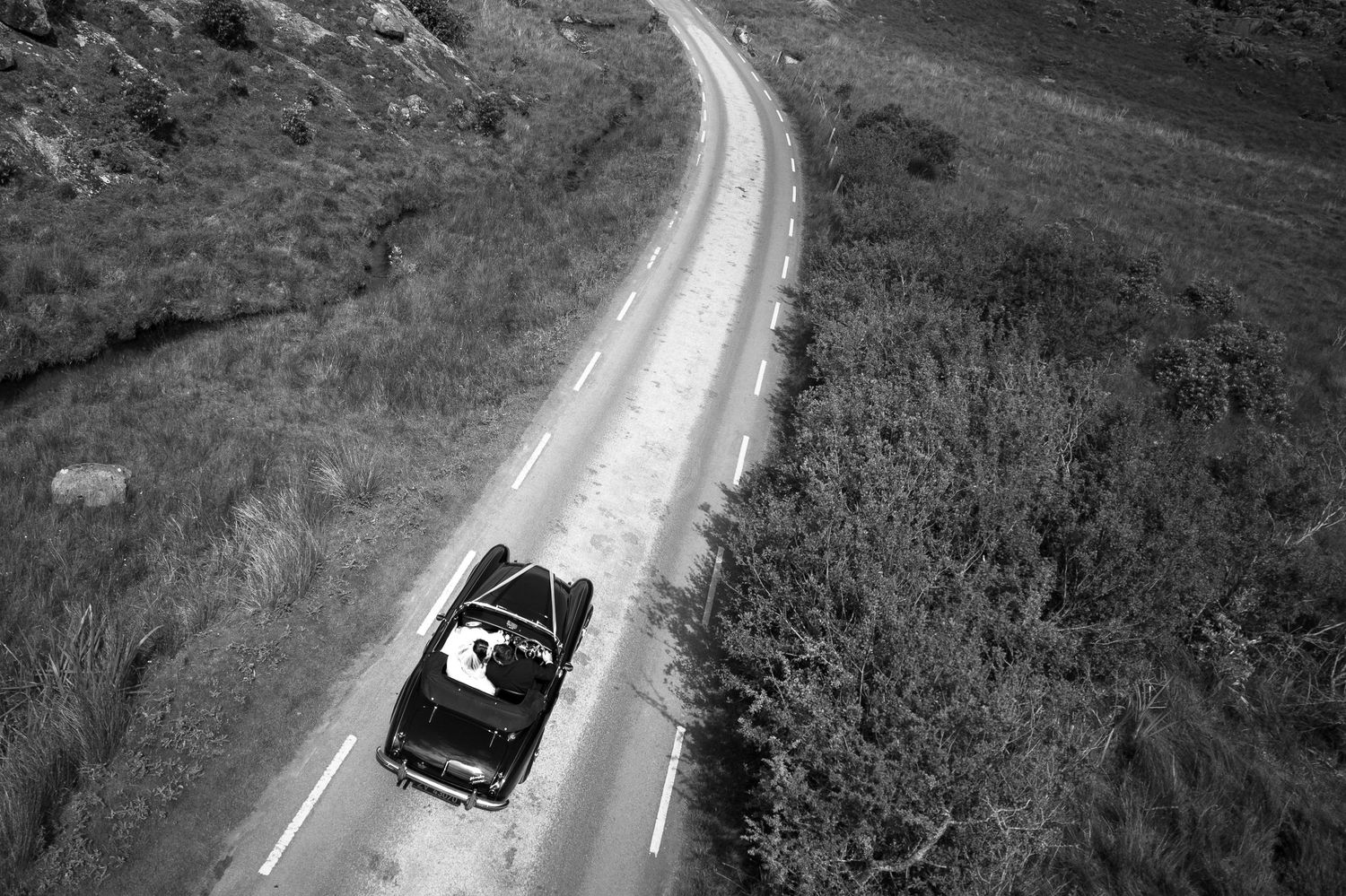 A black vintage car drives along a winding mountain road through a dramatic landscape in black and white.