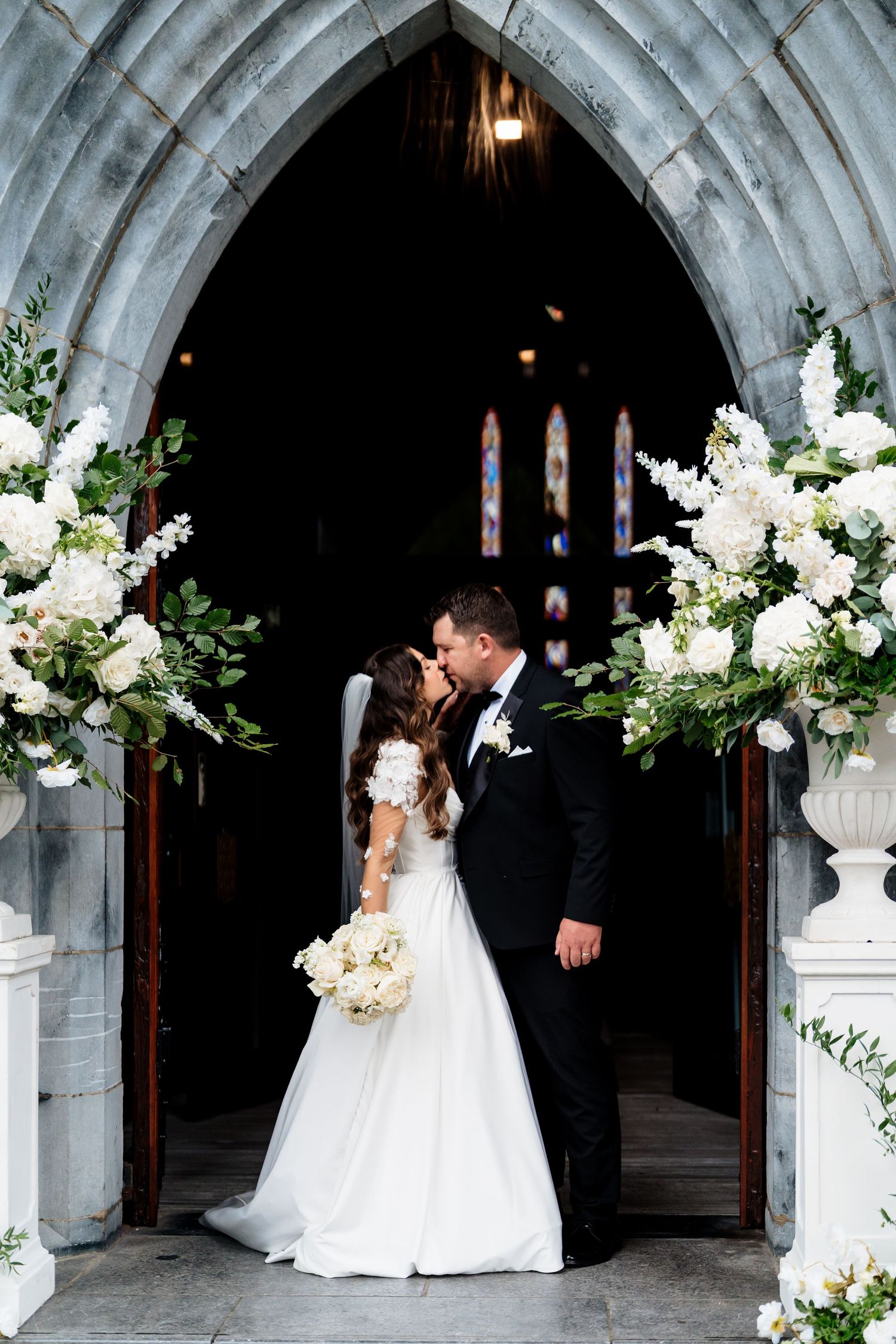 Elegant wedding couple poses under stone archway decorated with white floral arrangements and romantic lighting.