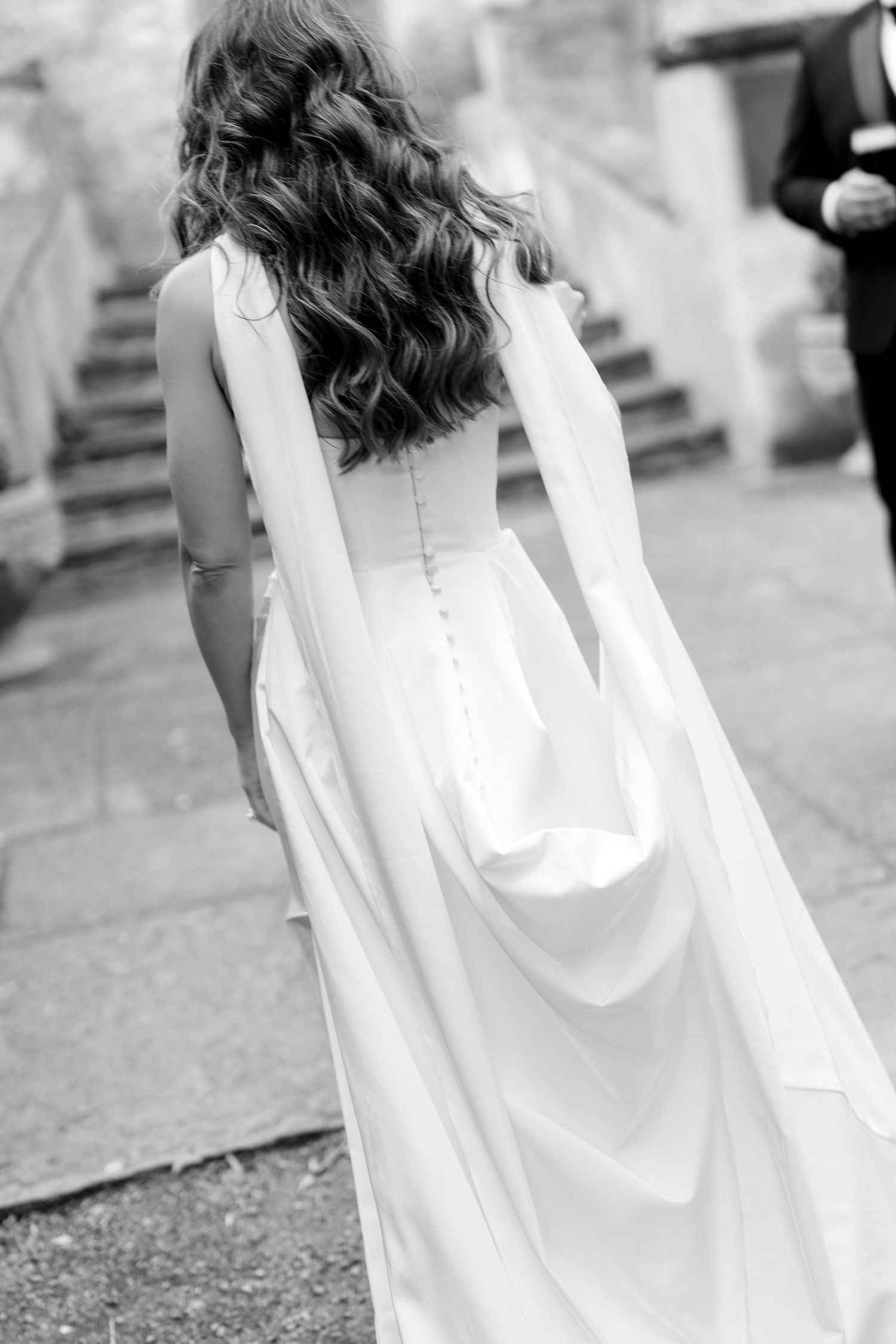 Black and white photo of bride in elegant wedding dress with flowing cape and long wavy hair seen from behind.