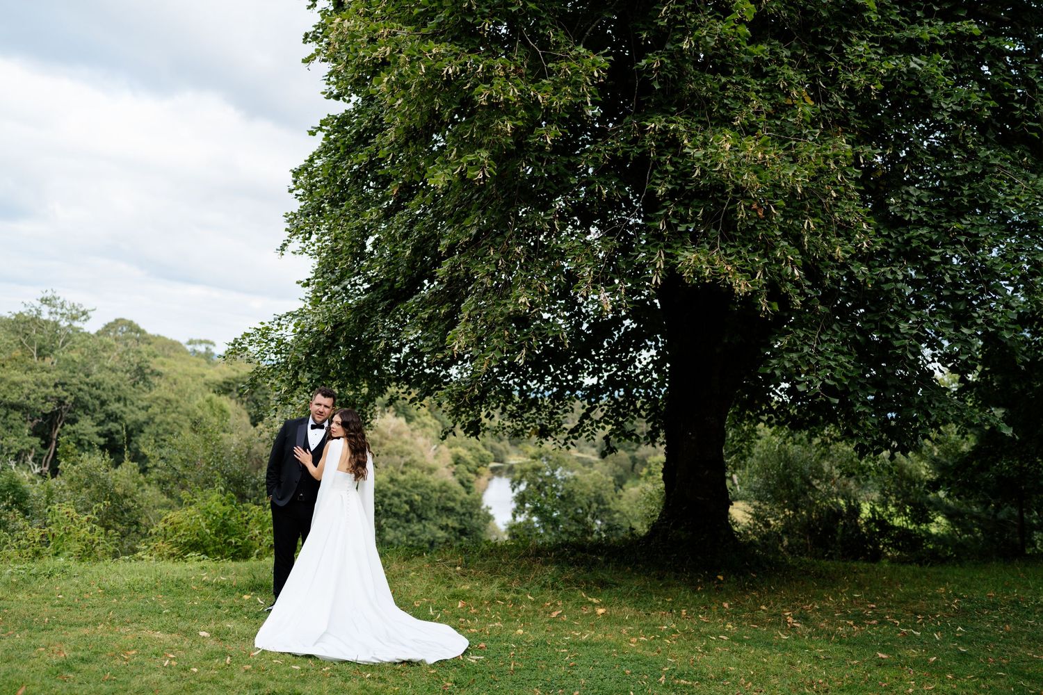 A couple in wedding attire stands together at a scenic outdoor location surrounded by lush green trees and foliage.