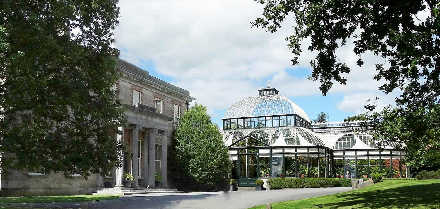 Historic stone building and glass conservatory structure in a manicured garden setting.