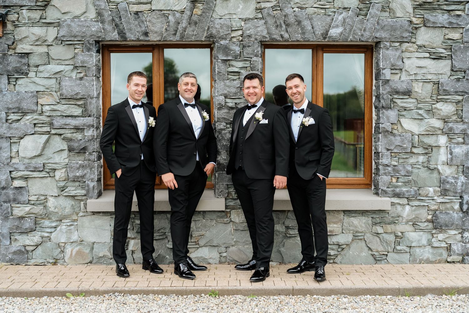 A group in formal black tuxedos sit with their feet propped up against a rustic stone wall.