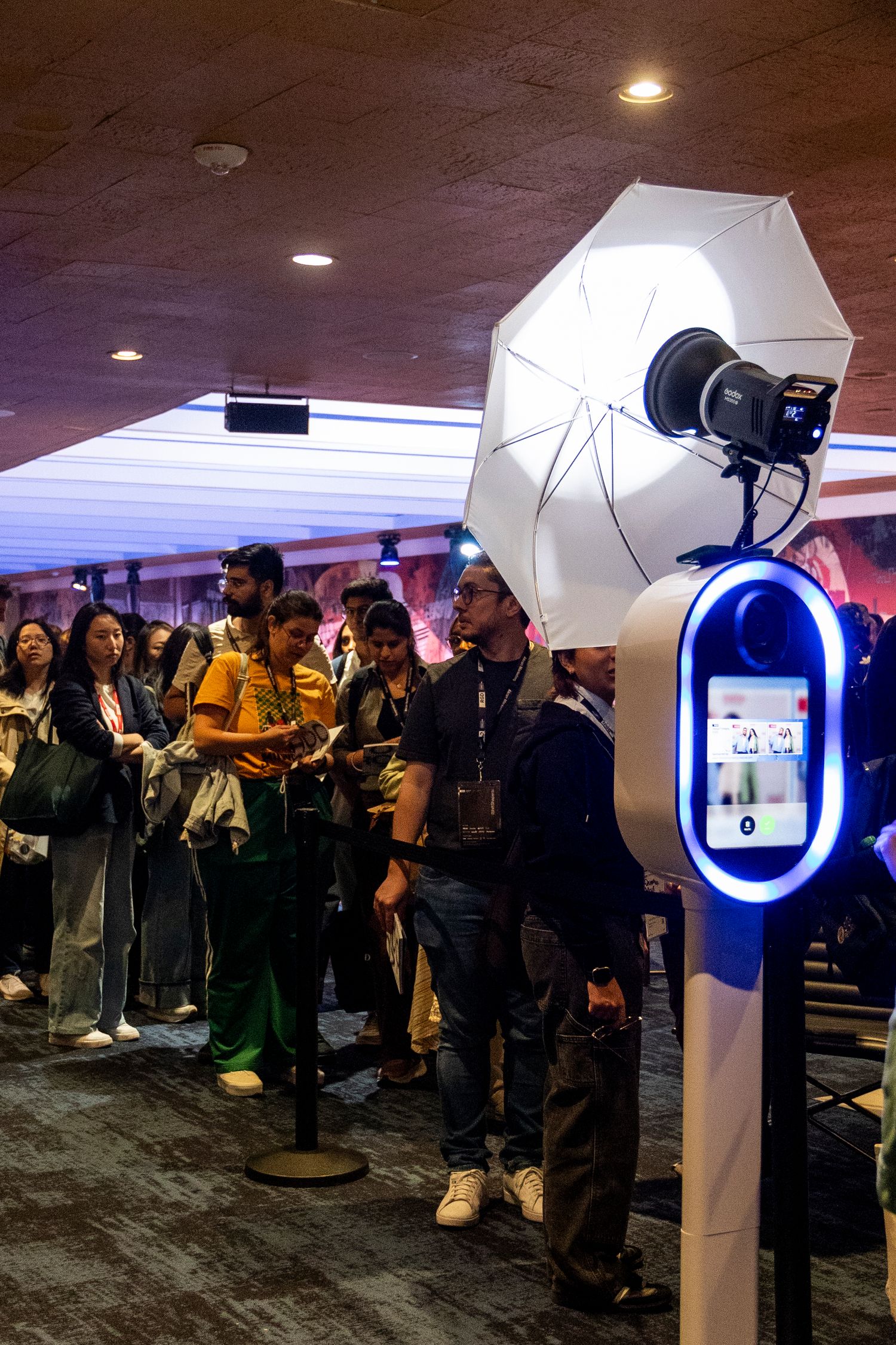 A crowd of people lined up at an event next to a photo booth station with a white umbrella light and blue kiosk.