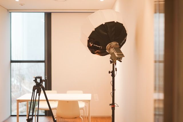 Professional photography studio setup with softbox light, tripod, and white table near large windows.