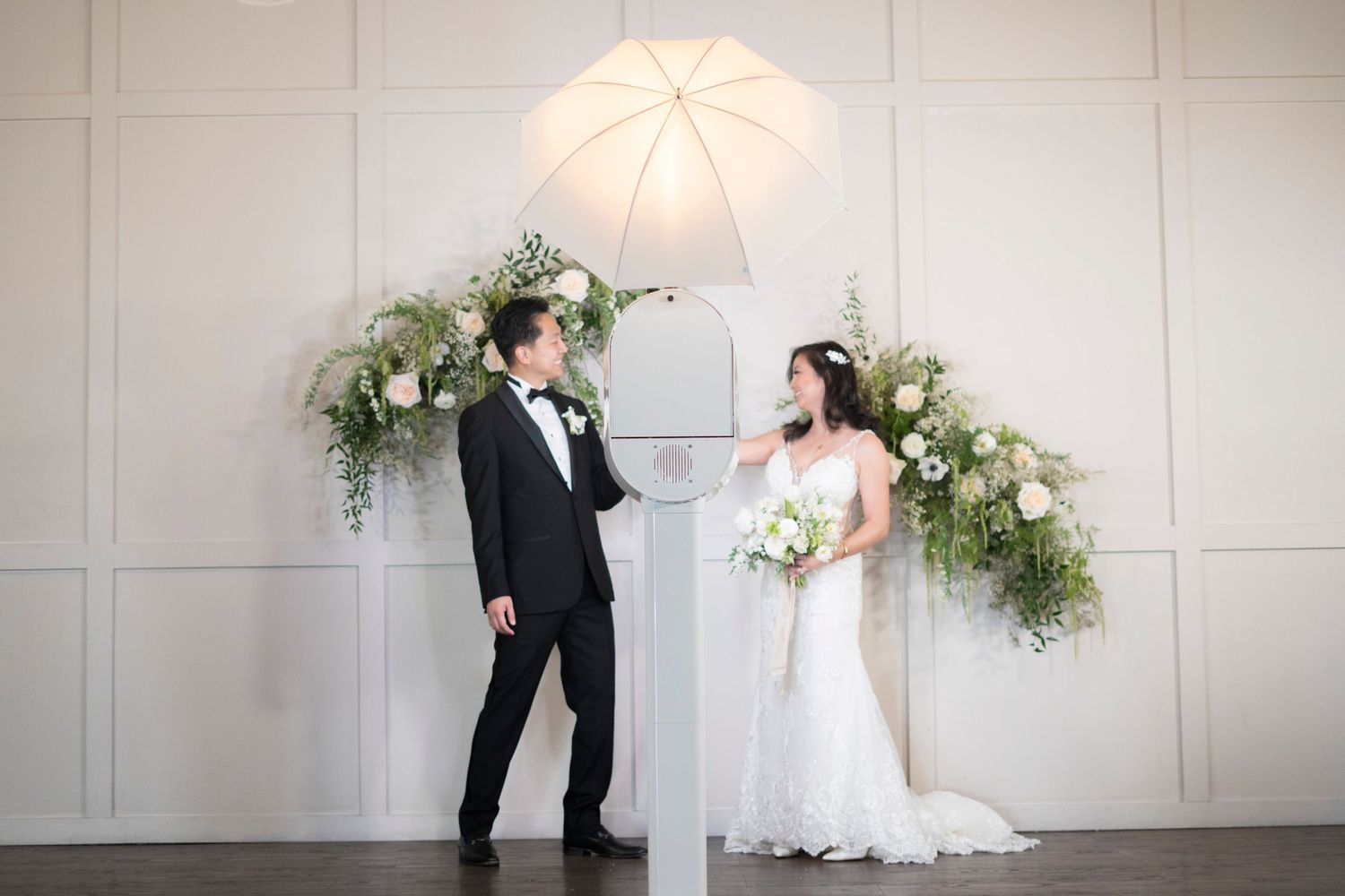 Wedding couple poses with white umbrella and floral arrangements against neutral wall.