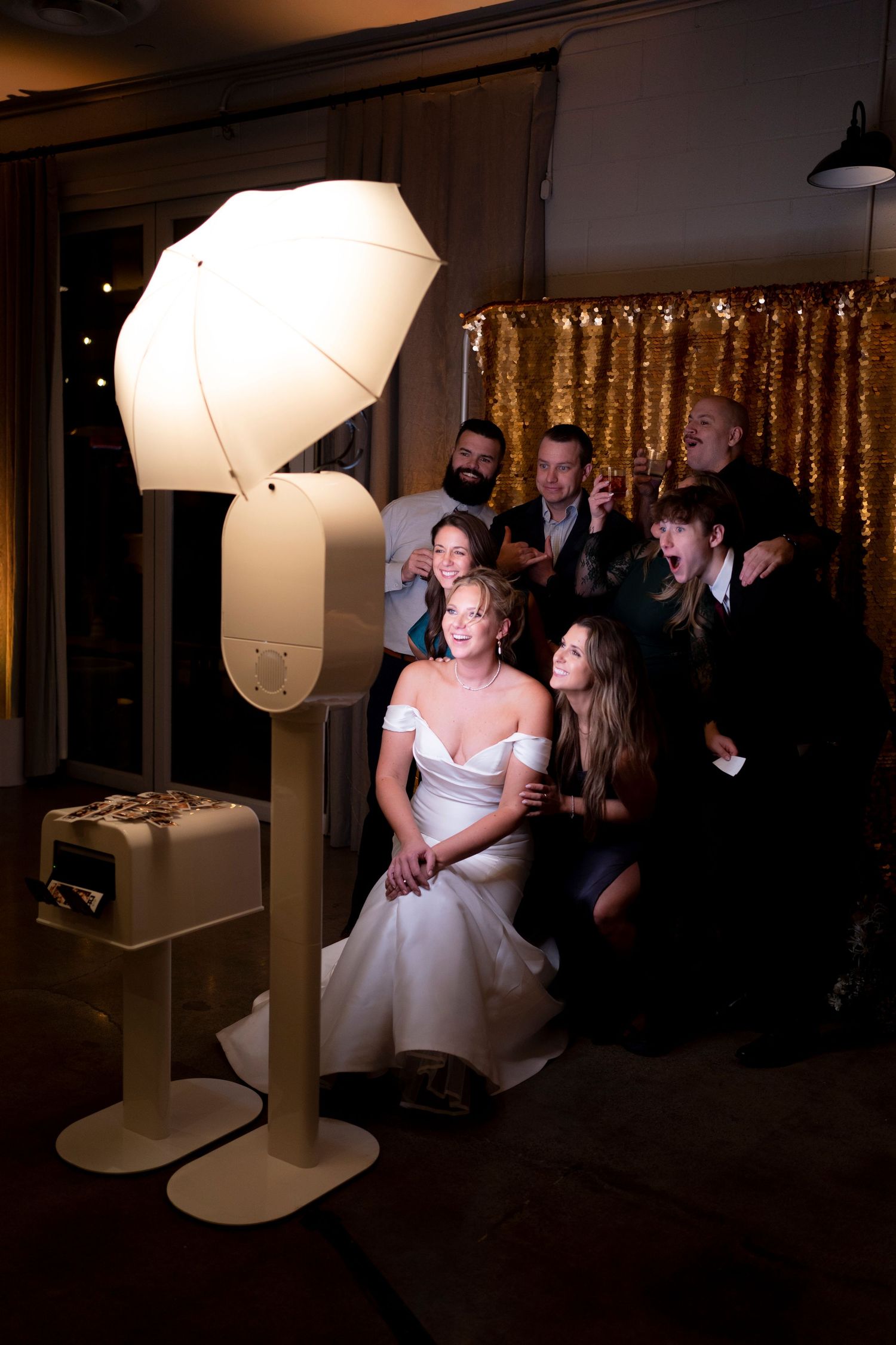 Bride in white gown poses with wedding guests at a photo booth with umbrella light against gold sequin backdrop.
