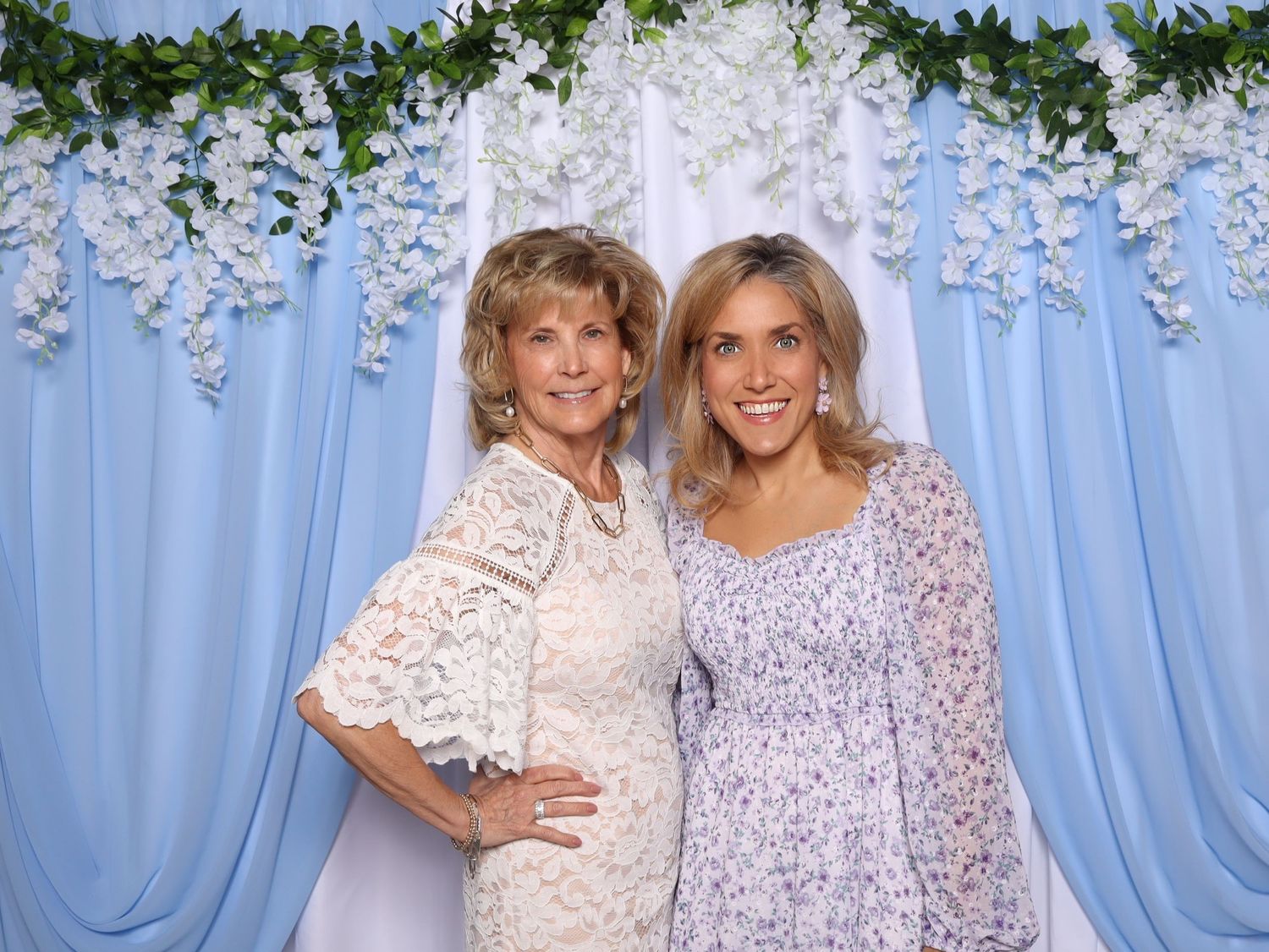 Two people wearing lace dresses pose together in front of a blue backdrop with hanging floral decorations.