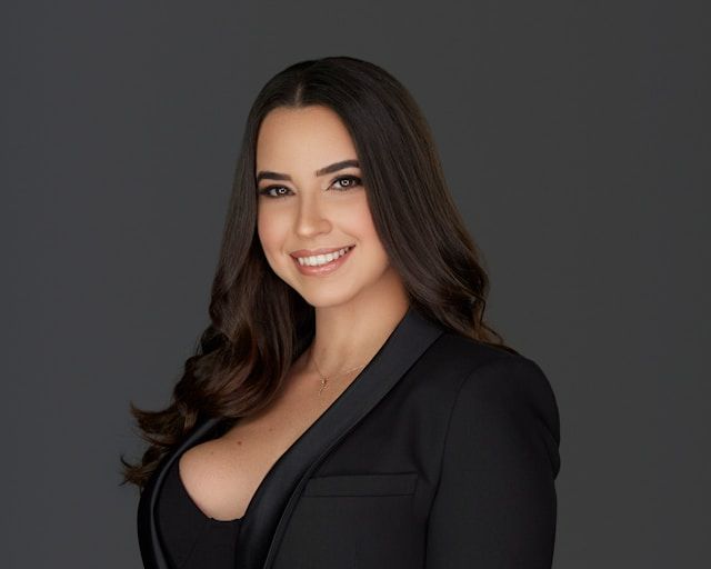 Professional woman in black blazer smiling confidently against gray background, corporate headshot photo.