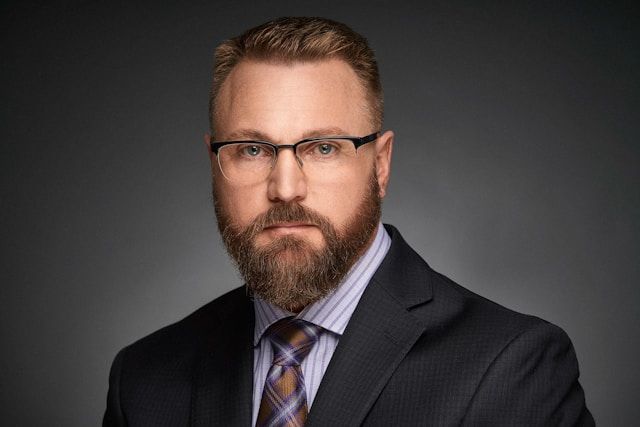 Professional headshot of a bearded man wearing glasses and a dark suit with a striped tie.