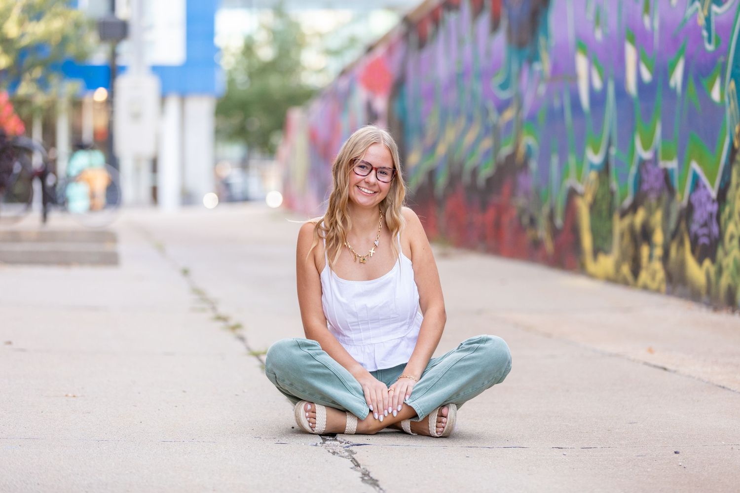 A high school senior with glasses and a white tank top sits cross-legged in front of a vibrant graffiti covered wall in Des Moines, Iowa.