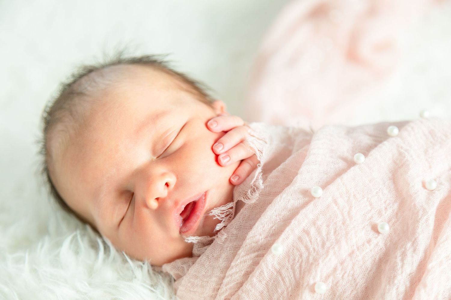 A newborn baby sleeps peacefully wrapped in a soft pink blanket, resting on a fluffy surface. The baby's hand gently touches their cheek.