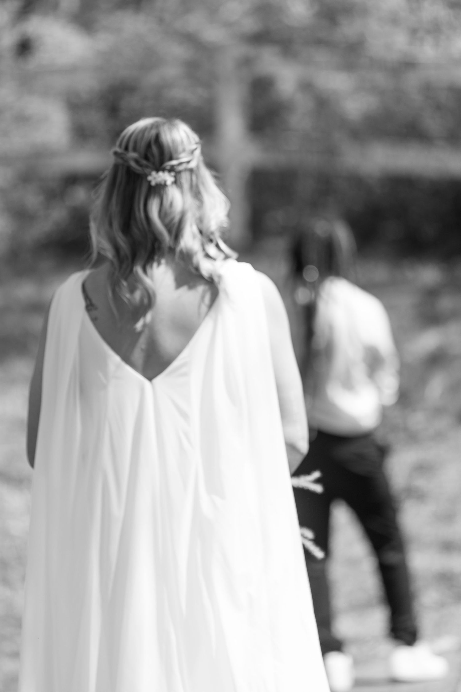 A black and white photo shows a woman in a flowing wedding dress with her back to the camera, looking towards her future husband.