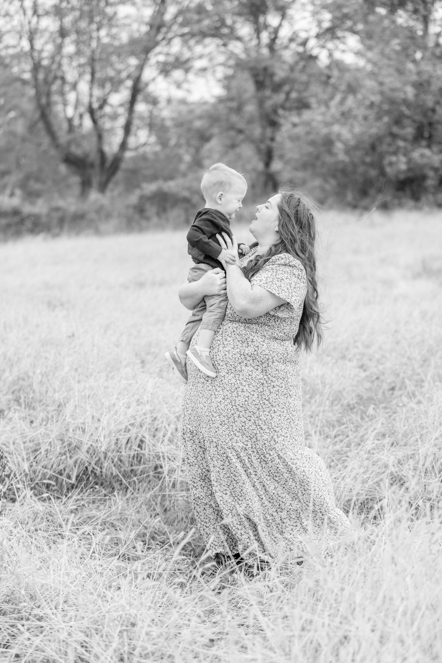 A mom in a flowing dress joyfully lifts her son in a grassy field. Trees in the background create a serene, affectionate atmosphere.