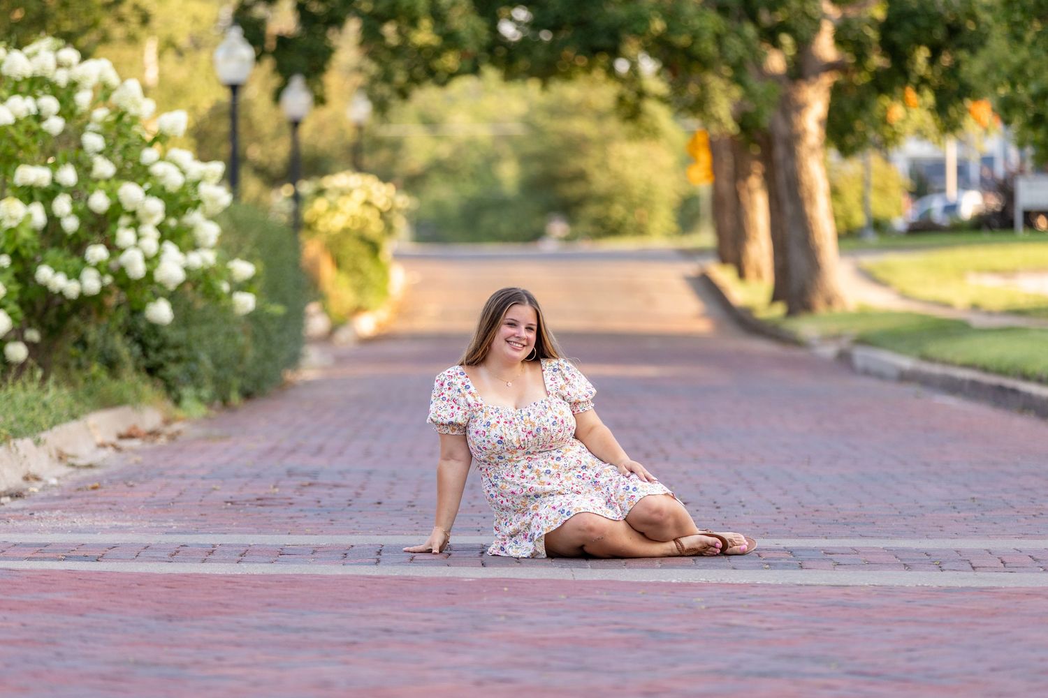 A high school senior in a floral dress sits on a brick road in Adel, Iowa under tree-lined street with soft, warm lighting.
