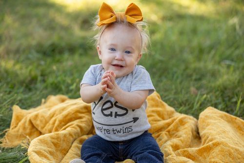 A smiling baby with a yellow bow sits on an orange blanket in a grassy area. The baby wears a gray t-shirt and jeans, conveying joy and play