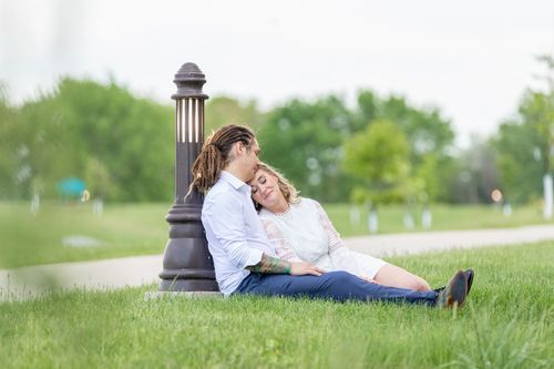 A couple sits on grass, leaning against a lamppost. They are relaxed and content, with the woman resting her head on the man's shoulder.