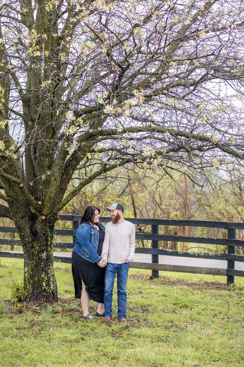 A couple stands under a blooming tree, holding hands and smiling at each other in a grassy field. A wooden fence lines the background, creat