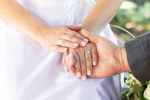 A close-up of a bride and groom's hands; bride's hands show a wedding ring, gently held by groom's hands. The tone is tender and intimate.