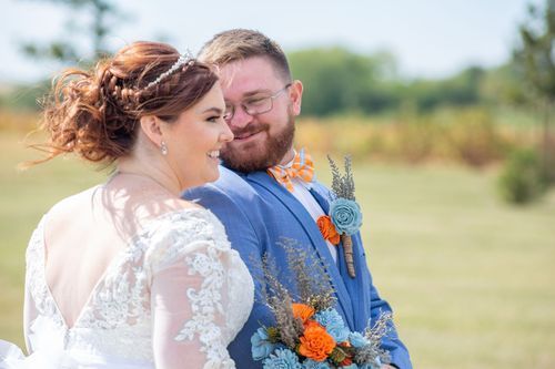 Bride and groom smiling outdoors at a sunny wedding. The bride wears a lace dress and tiara; the groom is in a blue suit with bright floral