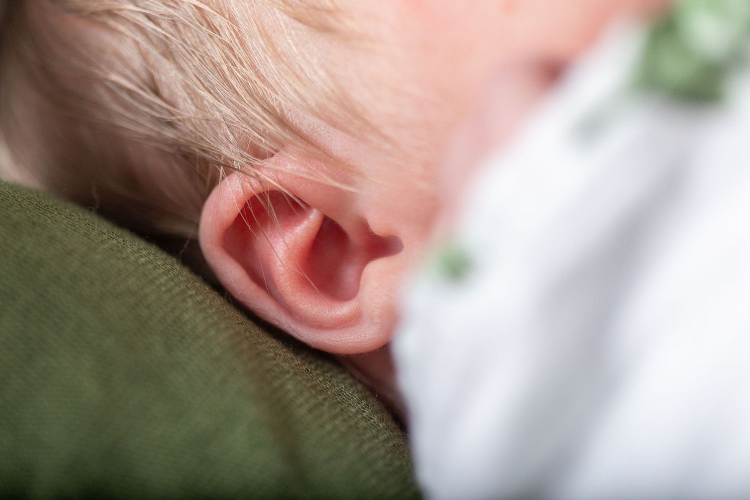 Close-up of a baby's ear with soft, blond hair, resting on a green blanket. The image conveys warmth and tranquility.