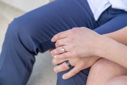 Hands with wedding bands resting on a knee, showing a diamond ring and black band. The setting suggests a relaxed, intimate moment.