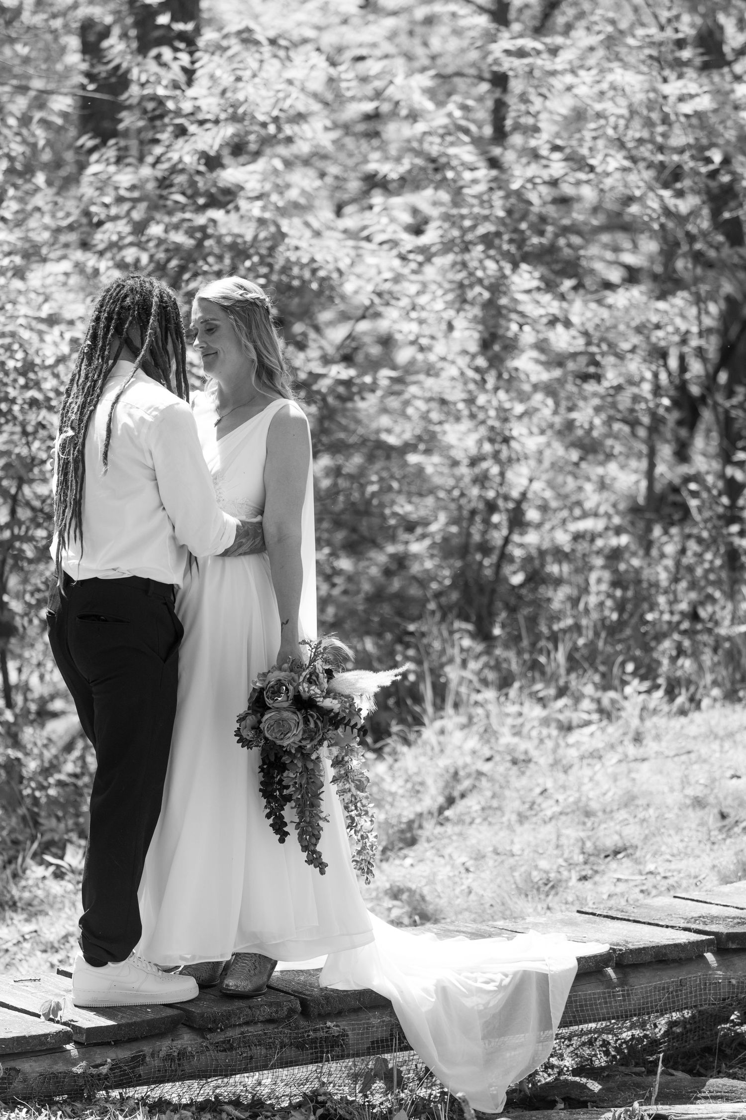 A couple stands on a wooden path in a sunlit, wooded area. The groom embraces the bride, who holds a bouquet.