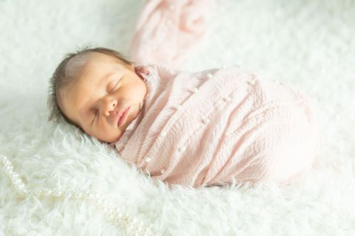 A sleeping baby wrapped in a pink blanket rests peacefully on a fluffy white surface. A delicate pearl necklace is draped beside the baby.