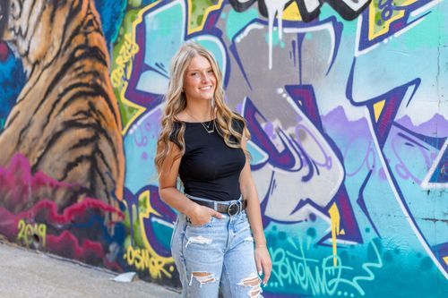 A high school senior in a black top and ripped jeans stands smiling in front of a vibrant graffiti mural in downtown Des Moines, Iowa.